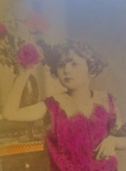 Una fotografía en blanco y negro de una niña con un vestido rosa brillante, sosteniendo flores, con un fondo antiguo que muestra una mesa decorativa.