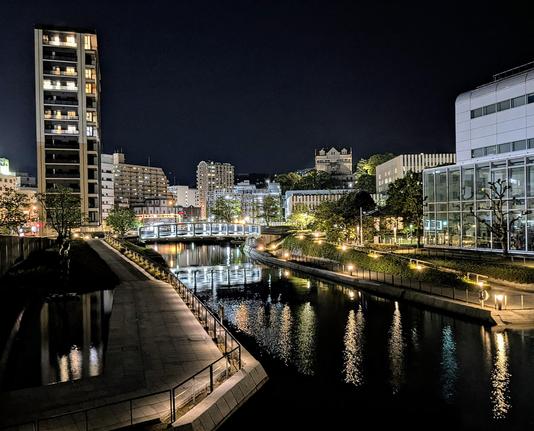 Illuminierte Uferpromenade. Die Licht spiegeln sich im Wasser.
Geländer und eine Brücke sind beleuchtet.