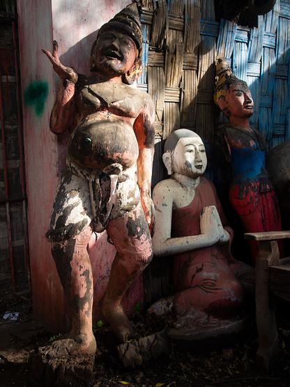 Three carved wooden buddha statues, all cracked with peeling paint, in front of a concrete wall with a woven rattan inset. Two are standing. The one closest has his right hand raised, palm up, possibly in the abhaya mudra, and is wearing a dodi. The center one is seated, has the most paint, white skin, and a red robe. He displays the namaskara mudra. The farthest is mostly obscured by shadow.