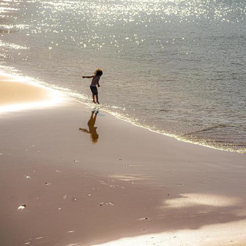 Child playing at the water's edge on a sandy beach. Sunlight reflects off the waves, creating a sparkling effect on the water. The child's silhouette and reflection are visible on the wet sand.