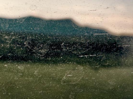 A blurred view through a dirty window, showcasing silhouettes of mountains in the background against a faded sky, with a greenish foreground.