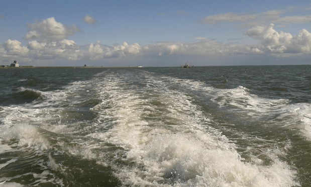Kielwasser eines großen Schiffs im blaugrünen Meer, am Horizont Land und kleine Schiffe, darüber blauer Himmel mit Wolken
