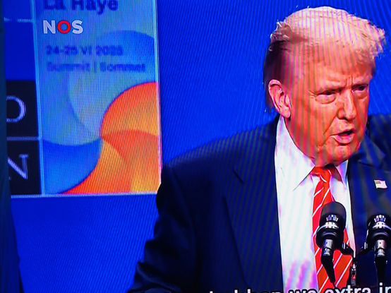 Foto van deel NOS live tv beeld met persconferentie: Donald Trump’s oranje gezicht naast oranje golf op NAVO-top poster op de muur erachter.