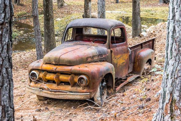 A severely rusted and weathered vintage pickup truck, likely from the 1940s or 1950s, sits abandoned in a wooded area. The truck's body shows extensive rust damage with patches of faded red, orange, and yellow paint still visible beneath the corrosion. The vehicle is missing its front bumper, headlights, and windshield, with the cab's windows empty. The truck bed appears intact but heavily deteriorated. It rests among tall trees with bare trunks, surrounded by fallen autumn leaves covering the forest floor. A small stream or water feature is visible in the background through the trees, and the lighting suggests it's daytime with natural forest lighting filtering through the canopy.