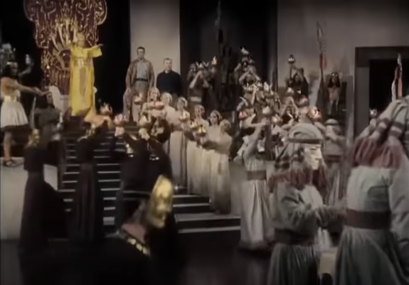 Scene from the old technicolor movie, some sort of bronze age style of ceremony with a woman dressed in yellow standing by the throne at top of short flight of stairs. The others are in white or black dresses, some wearing metalic masks. Two men wearing modern clothes are at honorary place next to the throne.
