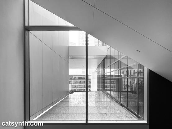 An angled ceiling cuts in front of a large glass window, through which we can see other steel and glass facades at various angles.