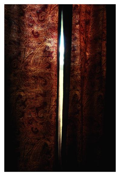 In a darkened room, a pair of red curtains is mostly drawn shut against the heat outside, but a narrow gap in the middle lets in some light. The paisley patterns on the curtain fabric are backlit by sunlight and glow against the shadows.