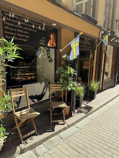 A charming cafe in Sweden with two wooden chairs on the sidewalk, Swedish flags, and a sign with a poem and portrait of Carl Michael Bellman. The cafe is named "Bellmans Cafe."