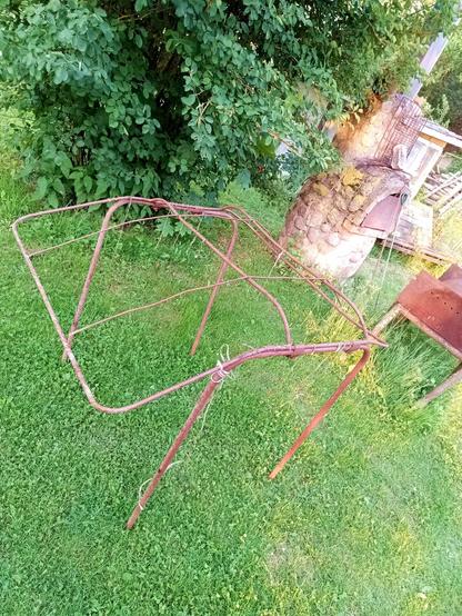 A bunch of rusty steel scrap sitting in the grass. It has four legs which can be folded in and a roof frame with a few thin bars between them. Some old strings dangle from the roof part. It could have supported some canvas and worked as removable roof to stick onto a tractor or other machine.