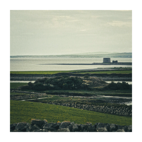 The photo is of a coastal landscape under a muted sky. In the foreground there is  green pasture land intersected by low, dry-stone walls.  In the mid-ground it shows a marshy area leading to a calm, grey-blue body of water.  In the far distance, a small, grey stone tower - Finvarra Martello tower , sits on a strip of land protruding into the sea.on the right hand side of the photo.