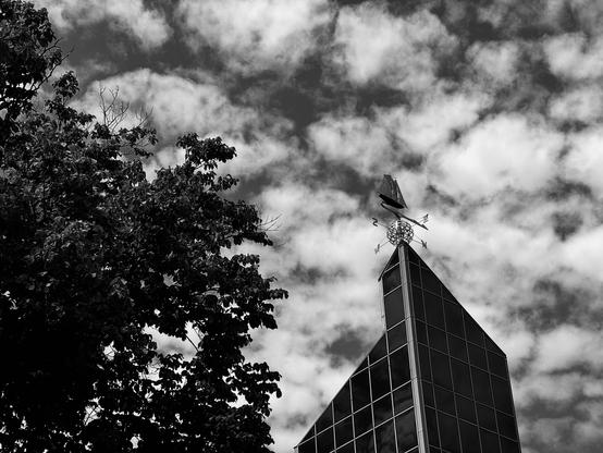A weather vane is seen on the top of an angular glass building against a party cloudy sky