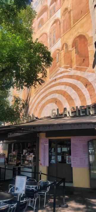 Mural encima de una heladería. El graffiti muestra un edificio con arcos y ventanas en colores tierra. Debajo se ve el letrero de la heladería y la puerta de entrada. Parte del graffiti está cubierto por un árbol.
