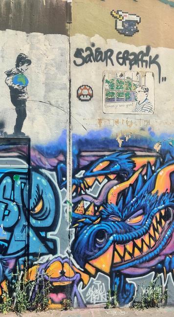 Graffiti en la Avd Debourg. Un niño con una bola del mundo en la mano, hace pis sobre un dragón azul. Hay diversas criaturas más en los mismos tonos de azul y amarillo que el dragón. En la parte superior hay un dos figuras de pixel art, una de un champiñón de Mario y otra de lo que parece una cabeza femenina. También hay un poster con un niño mirando por la ventana y pensando "Demain je serai grand"