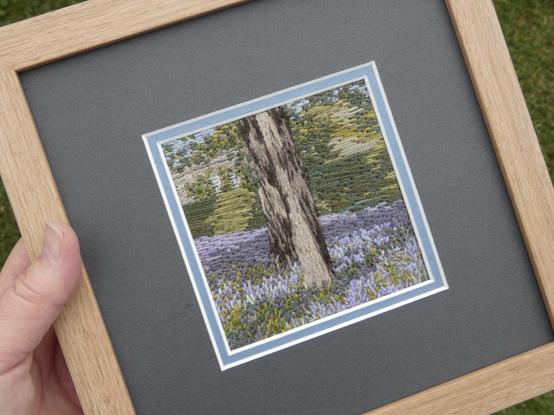 Stitched artwork depicting two tree trunks surrounded by bluebells. A hedge runs behind them and behind that can be glimpsed a corner of the Hall. Mounted in a pale blue and dark grey double mount and framed in abare oak frame 8x8 inches. Held in the artist's hand for scale