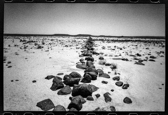 A black and white image of a line of stone in the sand. It barely looks like a “wall” but is part of one of the ancient animal traps known as “desert kites”.