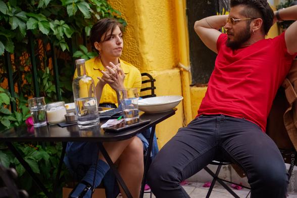 A candid outdoor café scene showing two people sitting at a small black table against a vibrant yellow wall with leafy green plants in the background. The table holds a glass water bottle, several glasses of water, a white bowl, a candle, a mobile phone, and a few small items. One person in a yellow shirt sits with hands clasped, while the other in a red shirt leans back with arms behind their head, creating a relaxed atmosphere.