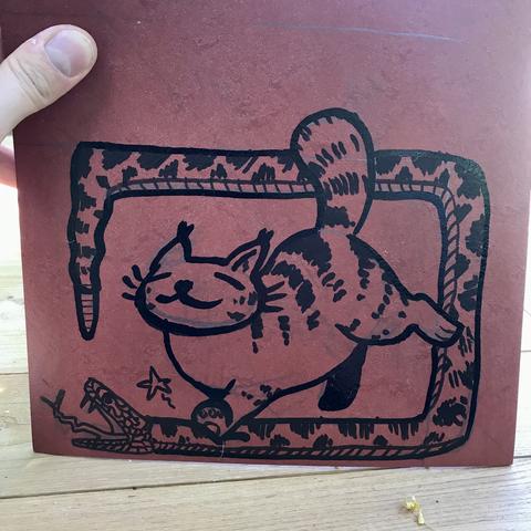 Dessin au feutre noir sur une plaque de lino marron : un gros chat rayé écrase un crotale. Le corps du serpent sert de cadre
