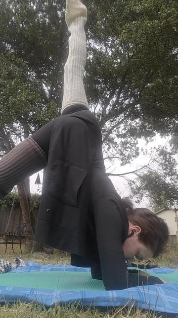 me (irene) doing a one-legged dolphin yoga pose outside in a houseyard. im wearing different colored legwarmers but the raised leg is pointing perfectly vertically. also "something something cakes in a bakery" bc my butt is looking very shapely today