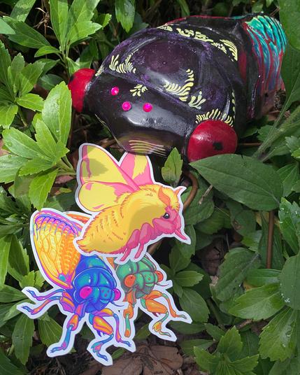 Photo of a large plaster cicada painted in bright colors "holding" some cicada stickers and a rosy maple moth sticker as it sits nestled in shrubbery