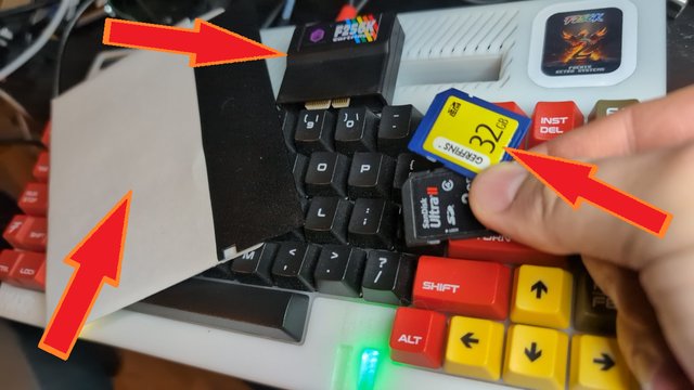 A F256K2 wedge case retro computer is seen under a 5.25" floppy disk in its sleeve, a flash retro style cartridge and a pair of SD memory cards held by a hand.