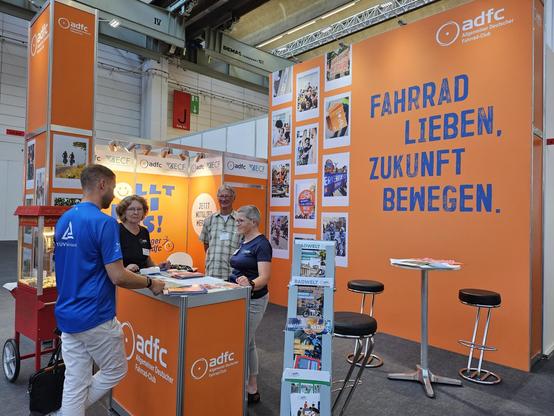 Messestand des ADFC mit orangenen Wänden. An einem Tisch stehen vier Personen, die sich unterhalten.