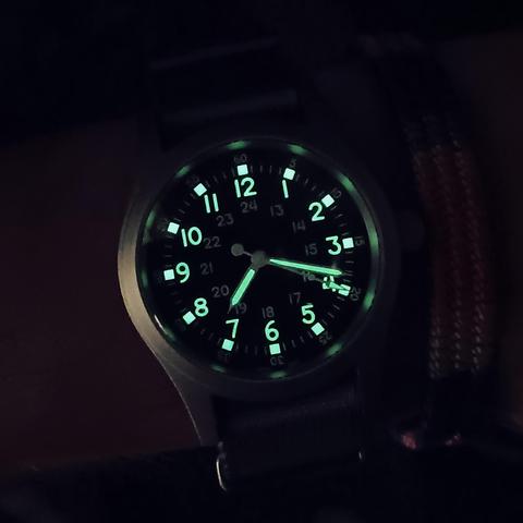 A watch with it's numerals, indices and hands glowing green in a darkened room.