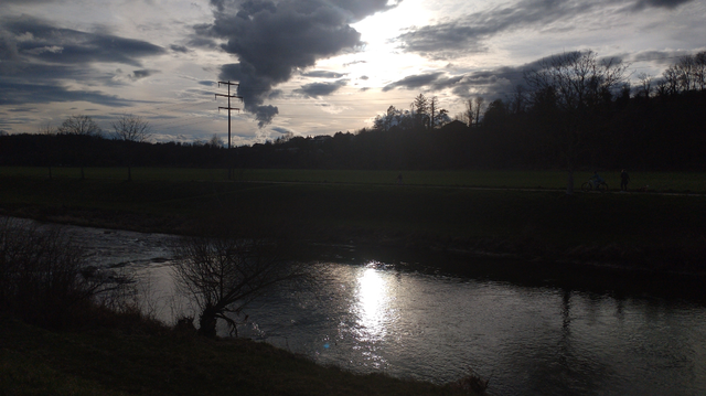 Blick über die begradigte Wutach, am Himmel mehrere dunkle Wolkenstreifen, die Sonne teilweise hinter einem davon verborgen. Sie spiegelt sich trotzdem im Flusswasser. Insgesamt eher monochromes Bild mit leichten Blau- und Gelbstichen.
