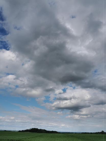 Cloud-filled sky with large, thick gray clouds dominating the upper part. A gap reveals patches of blue sky on the left. Below, a horizon with distant wind turbines peeks out between a stretch of green fields and a line of trees.