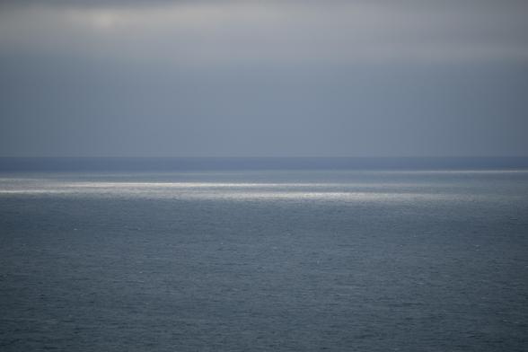 Blue-gray sea and gray overcast sky makes a dark image. The surface of the water is calm, just textured with small wavelets all the way out. 
At the top of the image there are some slightly lighter colored banks of cloud. On the surface of the sea, way out, light breaking through the clouds above is creating thin patches of white and silver light that stand out in contrast to the rest of the image.
