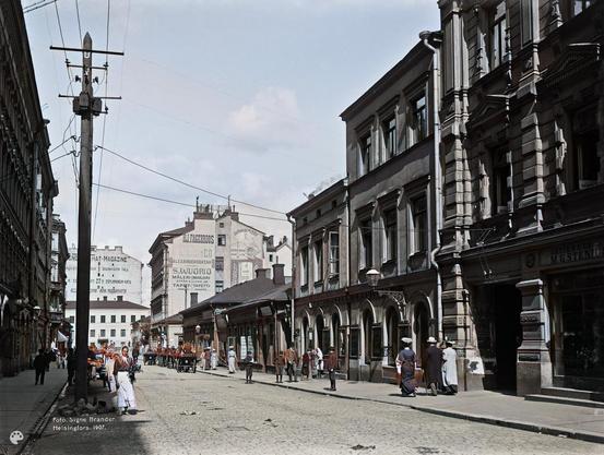 Kluuvikatu 3, 5, 7. (1907, Helsingin kaupunginmuseo)

Alkuperäisen valokuvan on ottanut Signe Brander. Tämä kuva on keinotekoisesti väritetty DeOldify-algoritmilla.