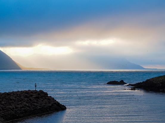 Dramatic, cloudy skies with some sun rays trying to get through, over a fjord with mountains in the background partly obscured by the ouds and a small harbour.
