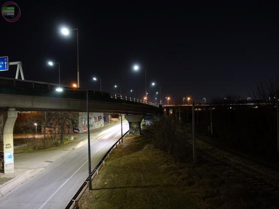 Autobahnhochstraße bei Nacht, darunter ebenfalls eine beleuchtete Straße, daneben im Finstern zwei Bahngleise