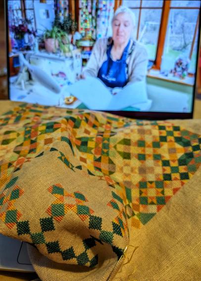 Unfinished multicolored geometric cross stitch embroidery project, while in the background a YouTube video is playing showing a grey haired woman behind a sewing machine.