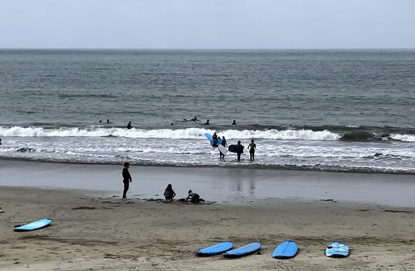 Ten or so surfers (or maybe some are paddle boarders) in the ocean, even though the waves are small. A couple people building a sand castle or some such on the beach. Five blue surfboards or paddle boards lie on the beach. It’s a gray day.