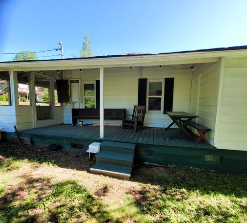 This is a photo of the exterior of a one-story, white house with a covered porch. The porch is made of dark green wood, and there are three steps leading up to it from the grassy yard.

On the porch, there's a dark wooden bench against the house wall to the left of the main window. To the right of the window, there's a dark wooden rocking chair and a picnic table with two benches. The house has two windows visible, both with dark shutters. String lights are draped across the porch ceiling.

The yard in front of the porch is covered in green grass with some patches of dirt and leaves, suggesting a natural, slightly unkempt look. The sky above is clear and blue, indicating a bright, sunny day. A utility pole and wires are visible in the upper left corner of the frame.