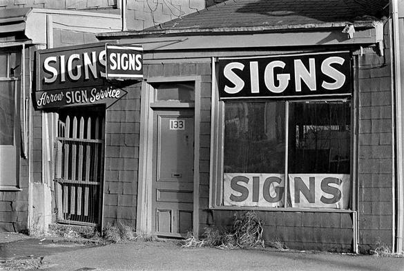 A photograph of an old timey store front, with multiple signs reading "Signs"