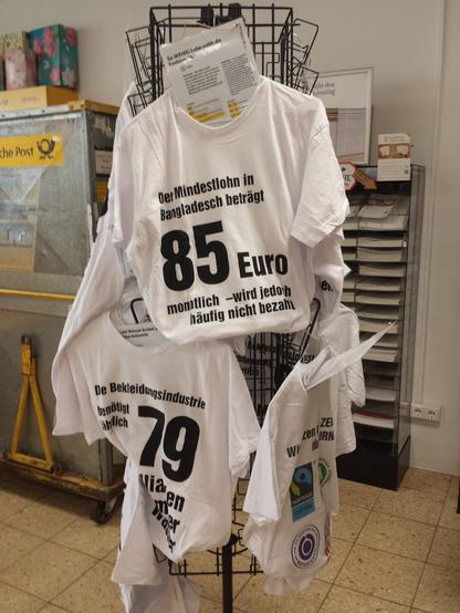 Ein Kartenständer wurde zweckentfremded: Es hängen mehree weiße T-Shirts mit verschiedenen Botschaften. Z.B. "Der Mindestlohn in Bagladesch beträgt 85 Euro monatlich - wird jedoch häufig nicht bezahlt". "Die Bekleidungsindustrie benötigt jedes Jahr 79 Millariden Liter Wasser". Ein T-Shirt zeigt wichtige Label, wie z.B. GOTS und FairTrade. Neben den Tshirts sind laminierte Din A4 Blätter mit weiteren Infos und Diagrammen. Im Hintergrund sind Artikel des Unverpackt Ladens, zB Schulhefte. Und links ist ein Transportwagen auf Rollen mit einem Deutsche Post Logo.
