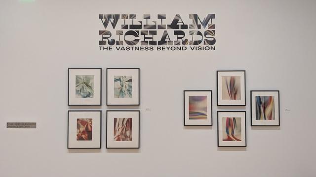 A white wall with abstract photographs grouped below a sign that reads William Richards The vastness beyond vision.