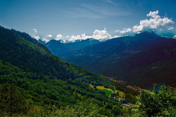 Panorama of a valley in the french alps during summertime. Both slopes of the valley are full of trees, there's a small town at the bottom. In the background, some mountains have snow.