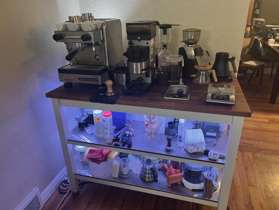 photo of an ikea kitchen island with seating setup as a coffee station with a faucet and sink including a pitcher/glass rinser, plumbed in commercial espresso machine, commercial espresso grinder, a home espresso grinder, a stagg pro kettle, bonavita scale (with hidden power), hario olive wood pour over set, hario siphon brewer. the shelves below have hidden led lighting showing off additional coffee equipment and supplies.  not visible is two 5 gallon water jugs hooked up to a flojet with an expansion tank and a 5 gallon jug for the drain lines in the area where chairs would be but in this case is against the wall