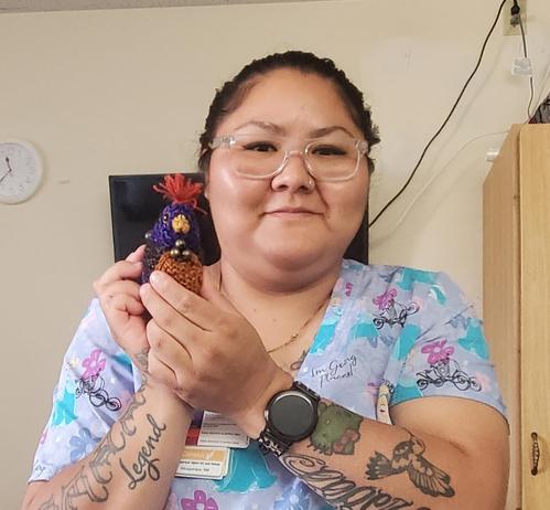 Medical aide,  a Navajo woman, her dark hair pulled back, in turquoise and pink scrubs, holding chickie up on her shoulder, revealing also her many  arm tattoos