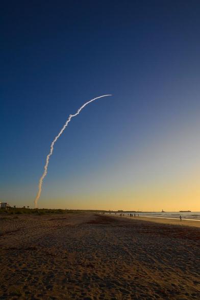 An Atlas V ascends on a column of smoke into the early morning sky. This is a very wide angle view that shows lots of sand on the beach, the ocean, a number of beach goers, and some buildings in the distance.