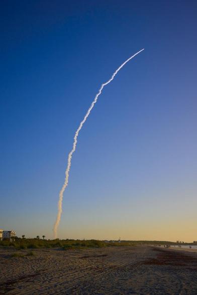 An Atlas V ascends on a column of smoke into the early morning sky. This is a wide angle view that shows sand on the beach, the ocean, a number of beach goers, and some buildings in the distance.