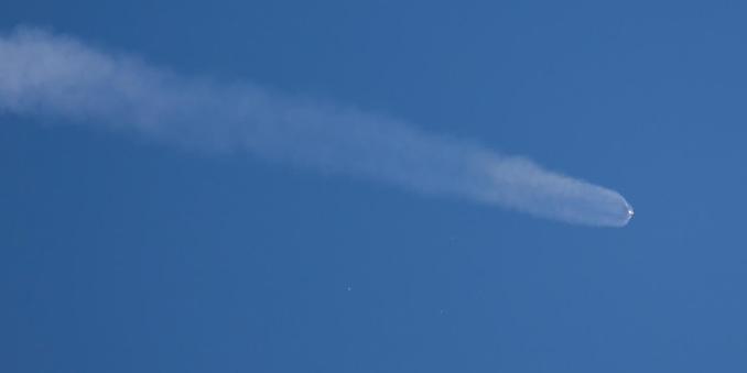An Atlas V of the 551 configuration shortly after the last two solid rocket boosters have separated from the first stage. Those boosters are still very close to the first stage. An exhaust plume trails the first stage through the blue sky. Below the plume are three small red-ish dots; these are the three boosters that separated earlier.