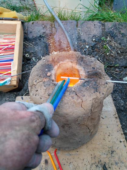 Eine kohlenstaubschwarze Hand hält gleichzeitig einen transparenten, einen grünen und einen blauen Glasstab über die Feuerdüse des Lehmofens. Eine orangene Flamme schlägt heraus.