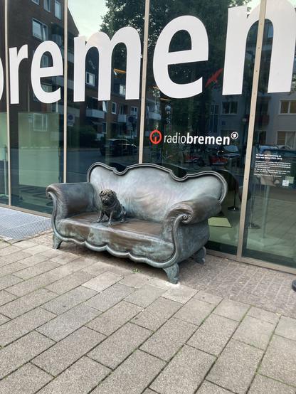 Scheibe mit Buchstaben "remen" und dem Logo von Radio Bremen, darunter ein Sofa, darauf ein Mops. Der Look ist Denkmal-artig.