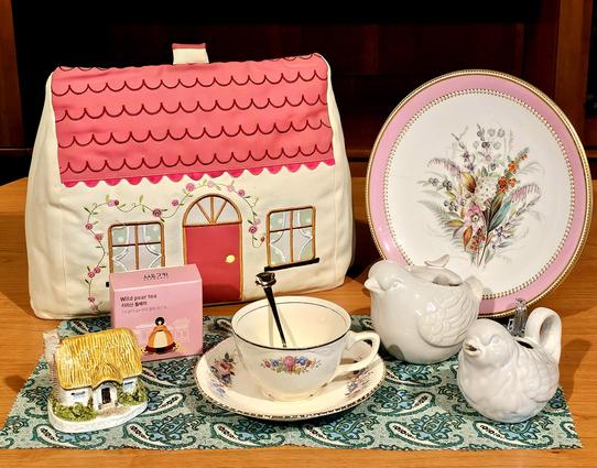 Nostalgic tea tableau in pastel colors with a vintage cottage theme. A large cottage-shaped tea cozy, a small thatched cottage figurine, china from Victorian and WWII eras, and figural bird cream and sugar, all on a paisley cloth. The tea is Wild Pear white tea.