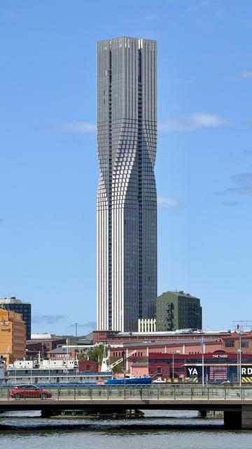 A tall, modern skyscraper with a twisting design is prominently featured against a blue sky. The building is flanked by lower urban structures, including red and green buildings, and there's a waterway with boats in the foreground.