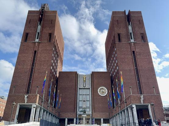 A large brick building featuring two towers and a central structure with sculptures. Rainbow flags are displayed along the facade, and a golden emblem is visible above the entrance. The sky is partly cloudy.
