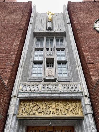 The image depicts a prominent architectural facade featuring a tall, decorative entrance with a golden statue atop. The building is made of gray stone with intricate carvings at the base, illustrating historical scenes and figures. Red brick walls flank the sides, and a clock.